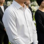 Osoba v bílé košili stojí mezi truchlícími na pohřbu s klidným pozadím hřbitova.