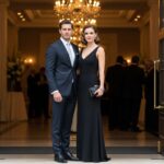 Muž a žena v elegantním oděvu stojí u vstupu na formální událost, pozadí s dekoracemi a hosty