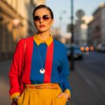 Stylová žena v barevném oblečení stojí na městské ulici za zlaté hodiny s brýlemi a šperky